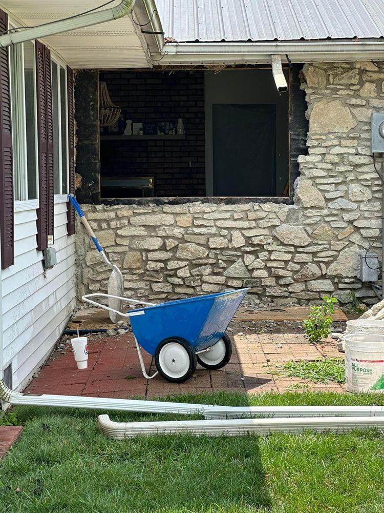 A blue wheelbarrow in front of a stone wall, open window, and a house with siding.