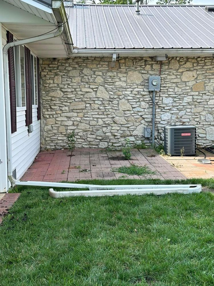 A white-sided house with a stone wall, metal roof, air conditioner, and a patio area with grass and discarded gutters.