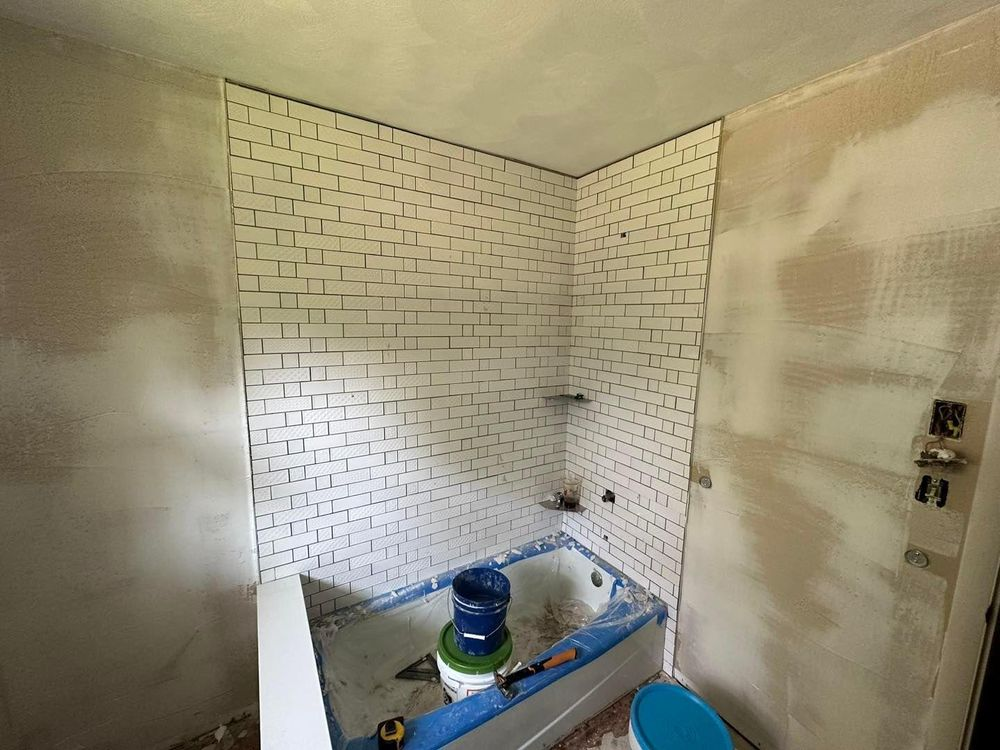 Bathroom renovation in progress: white brick-like tile on walls, unfinished plaster, bathtub, tools, and a blue bucket.