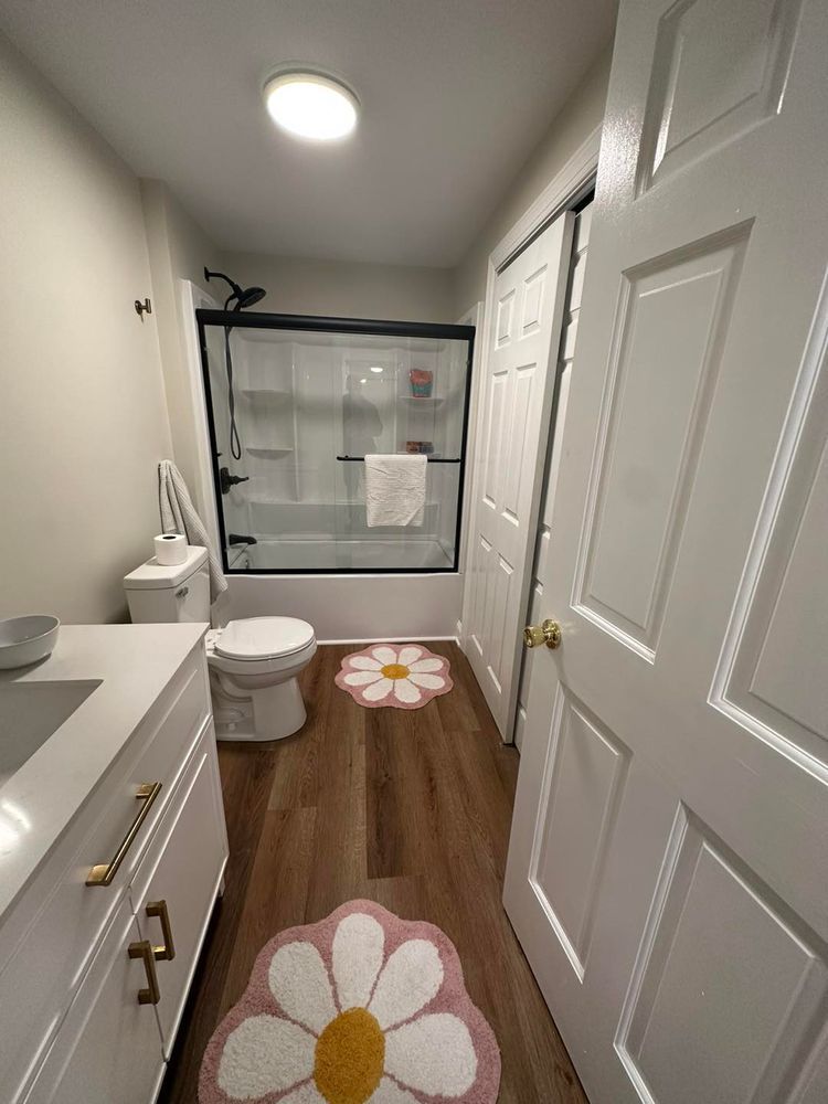Bathroom with white vanity, toilet, bathtub, and daisy-shaped rugs on wood-look floor.