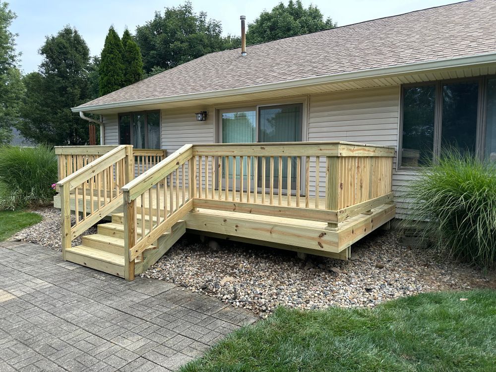Wooden deck with stairs and railings against a light tan house, set on a stone patio and grass lawn.