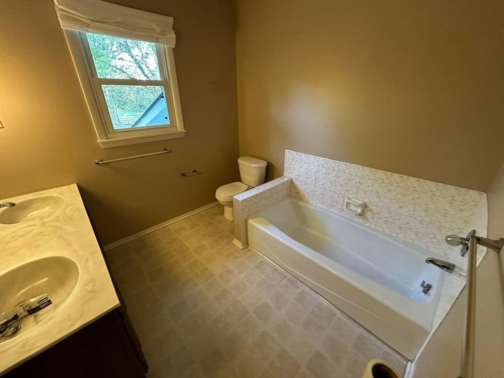Bathroom with a tub, toilet, and double sink; neutral colors, window, and towel racks.