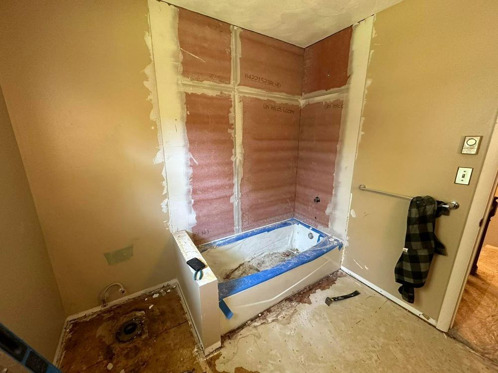 Bathroom renovation in progress: tub, shower walls, and surrounding walls exposed, with tools and debris on the floor.
