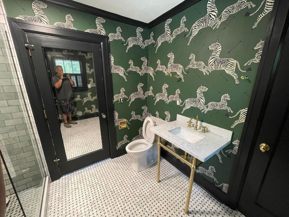 A bathroom with zebra-print wallpaper, black trim, marble sink, and white tile floor. A person is reflected in a large mirror.