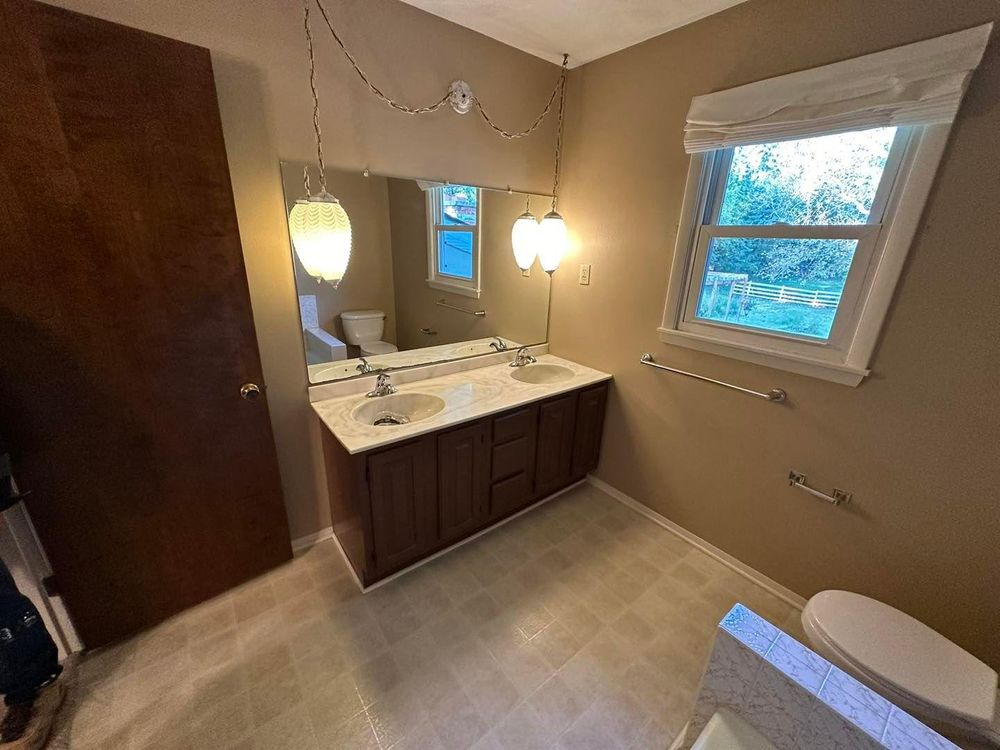 Bathroom with a vanity, mirror, window, and toilet. Brown cabinets, neutral walls. Two pendant lights.