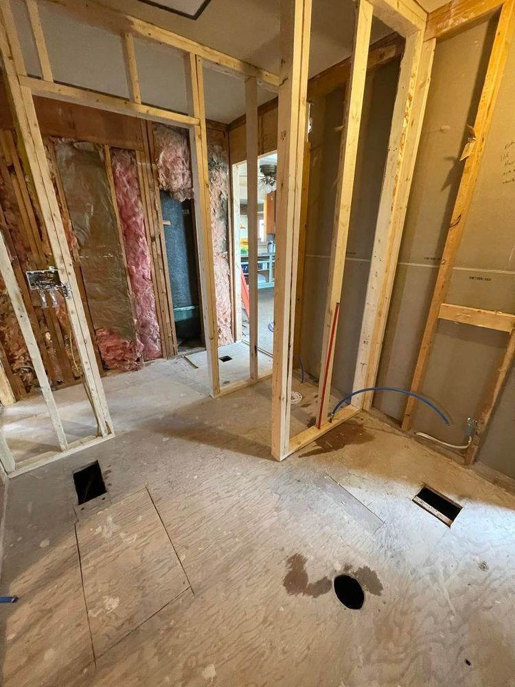 Bathroom under construction: bare wooden framing, exposed plumbing, subfloor, and insulation.