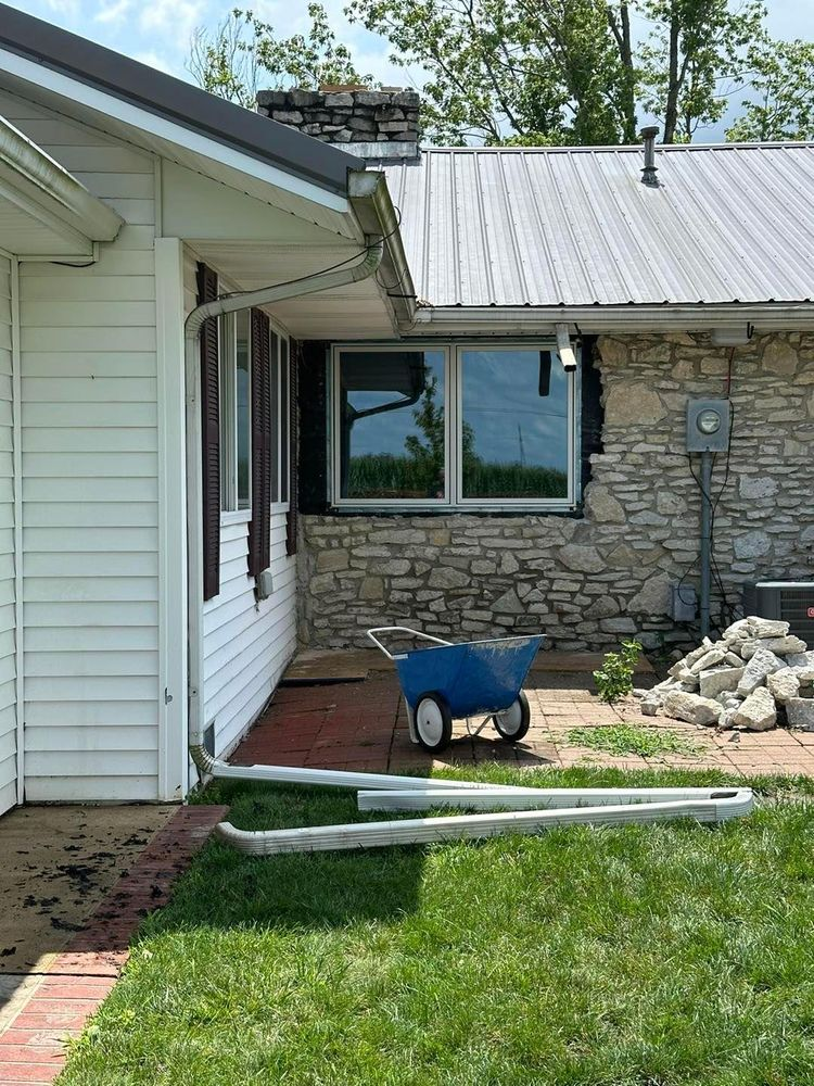 Exterior of a house with stone and siding, a wheelbarrow, and gutters on the ground.
