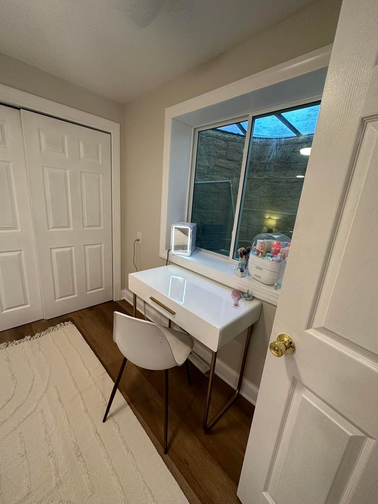 White vanity desk by a window in a room with a white door and rug, and a white chair.