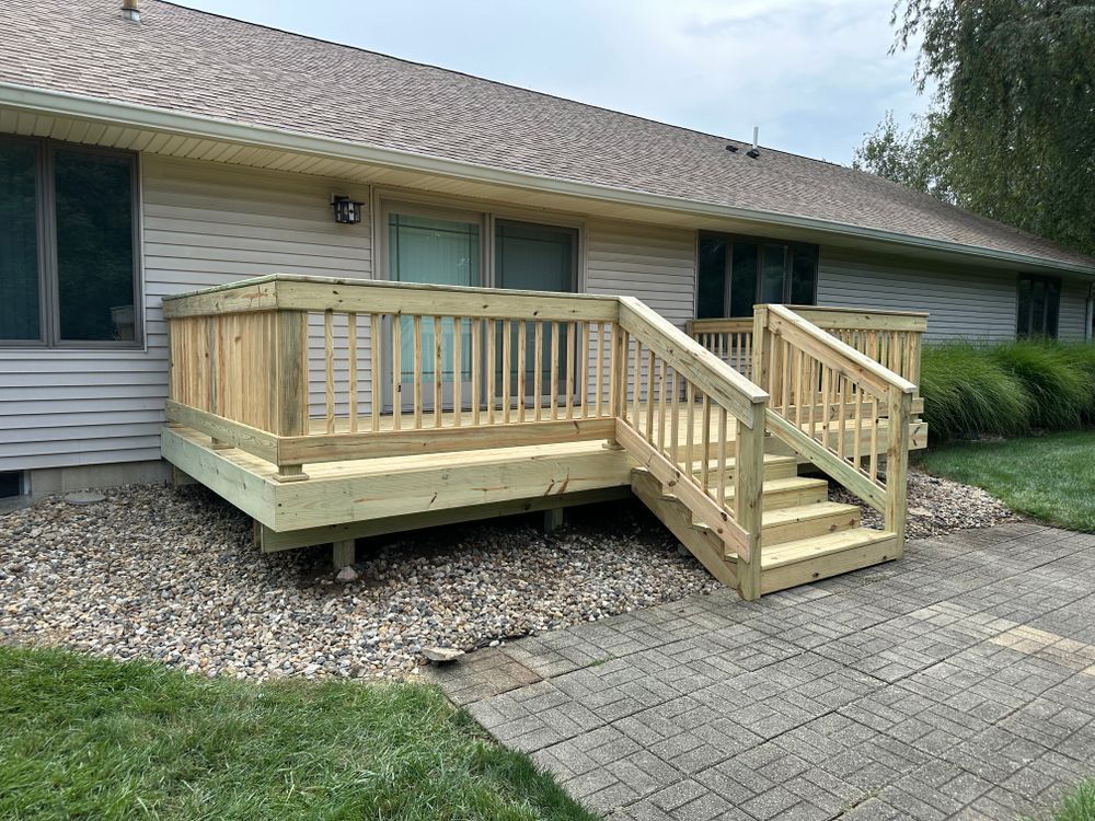 Wooden deck with stairs, next to a house on a stone patio, with grass and landscaping.