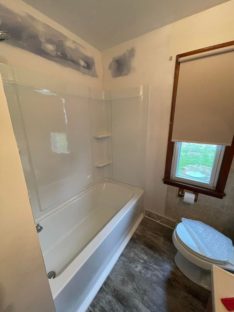 Bathroom with a white tub, shower surround, and toilet. The walls are painted. A window with a closed blind is on the right.