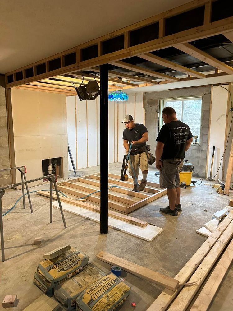 Two construction workers framing a room, surrounded by wood, cement bags, and tools.