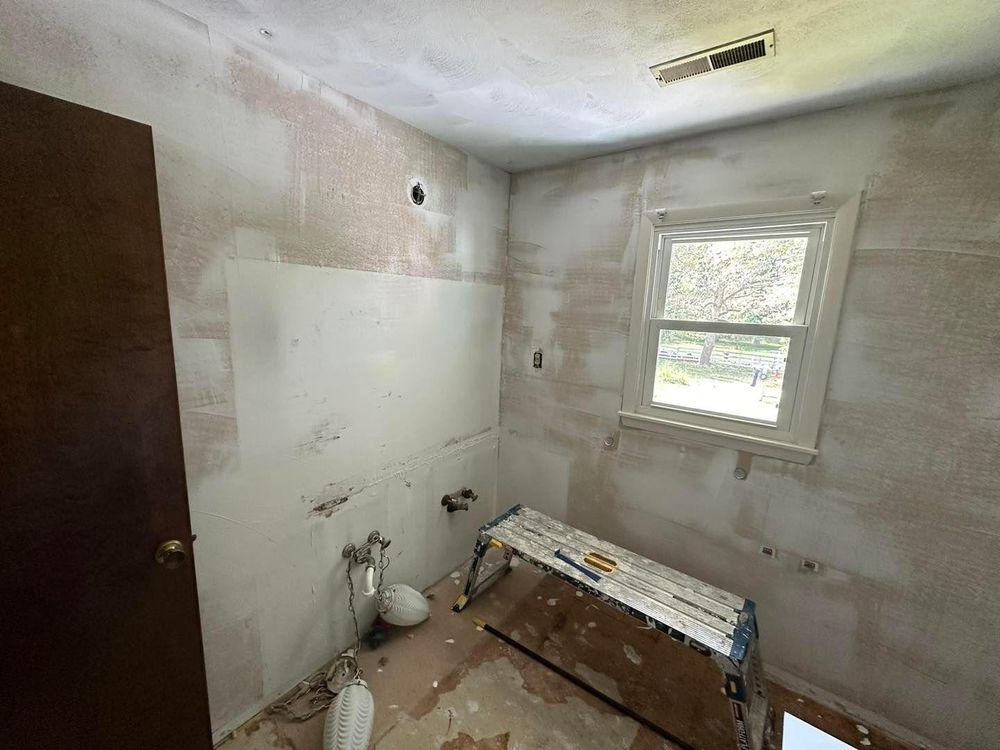 Bathroom under renovation; walls covered in primer, exposed plumbing. Wooden door, small window, scaffold.