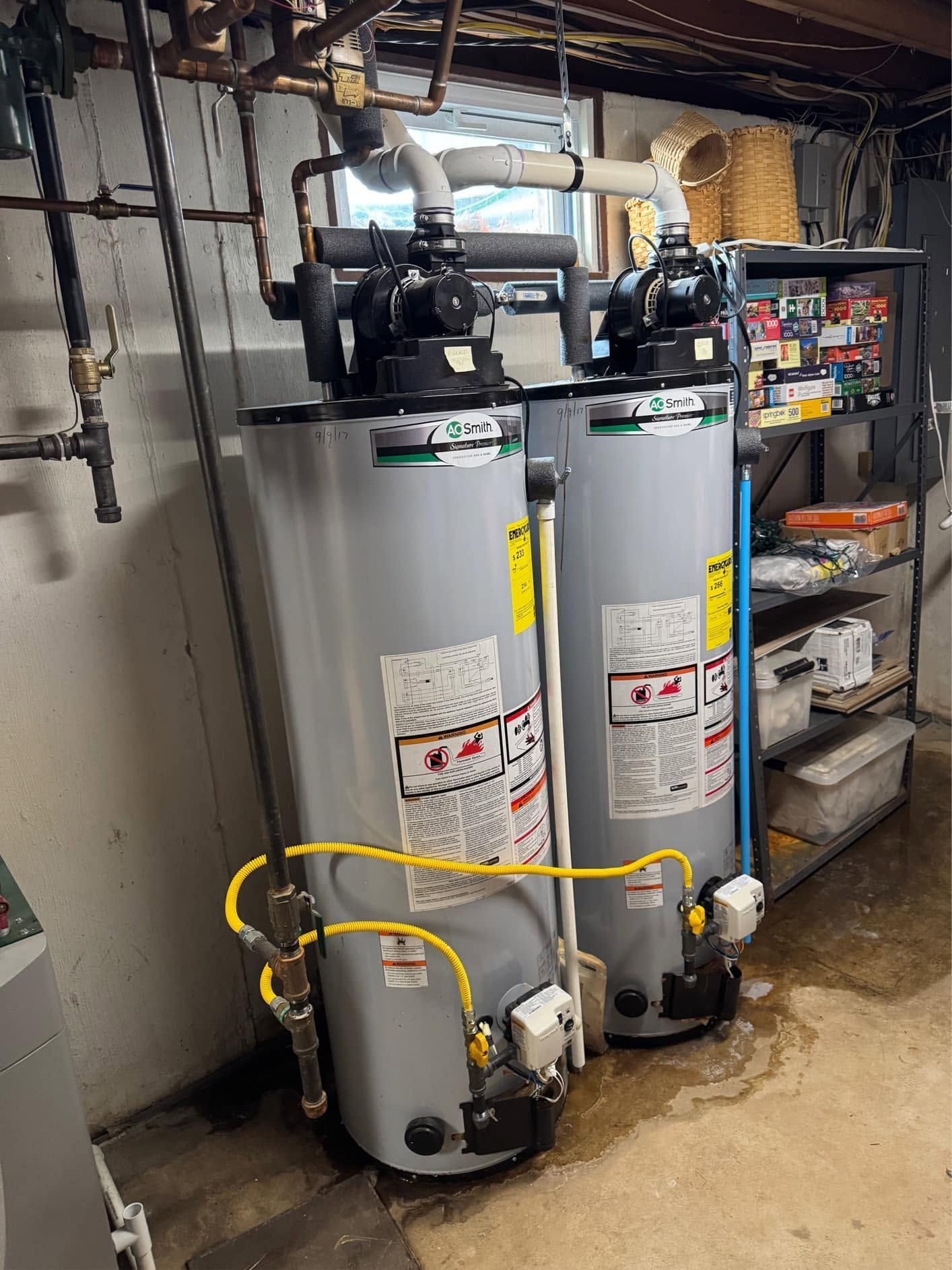 Two gray water heaters with pipes and yellow gas lines in a basement utility area