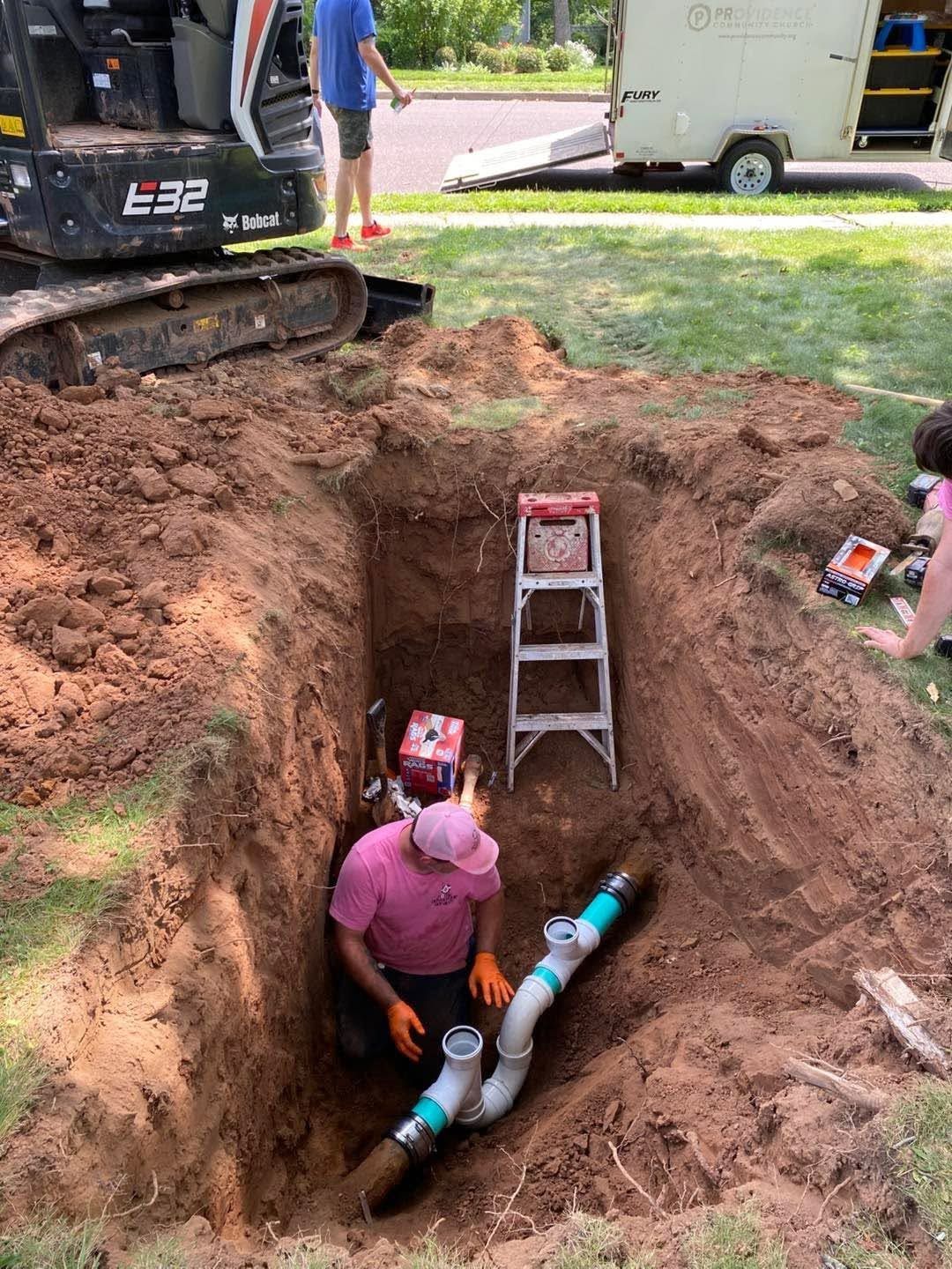 Worker in pink shirt repairing a broken water line in a deep trench beside a sidewalk and excavator.