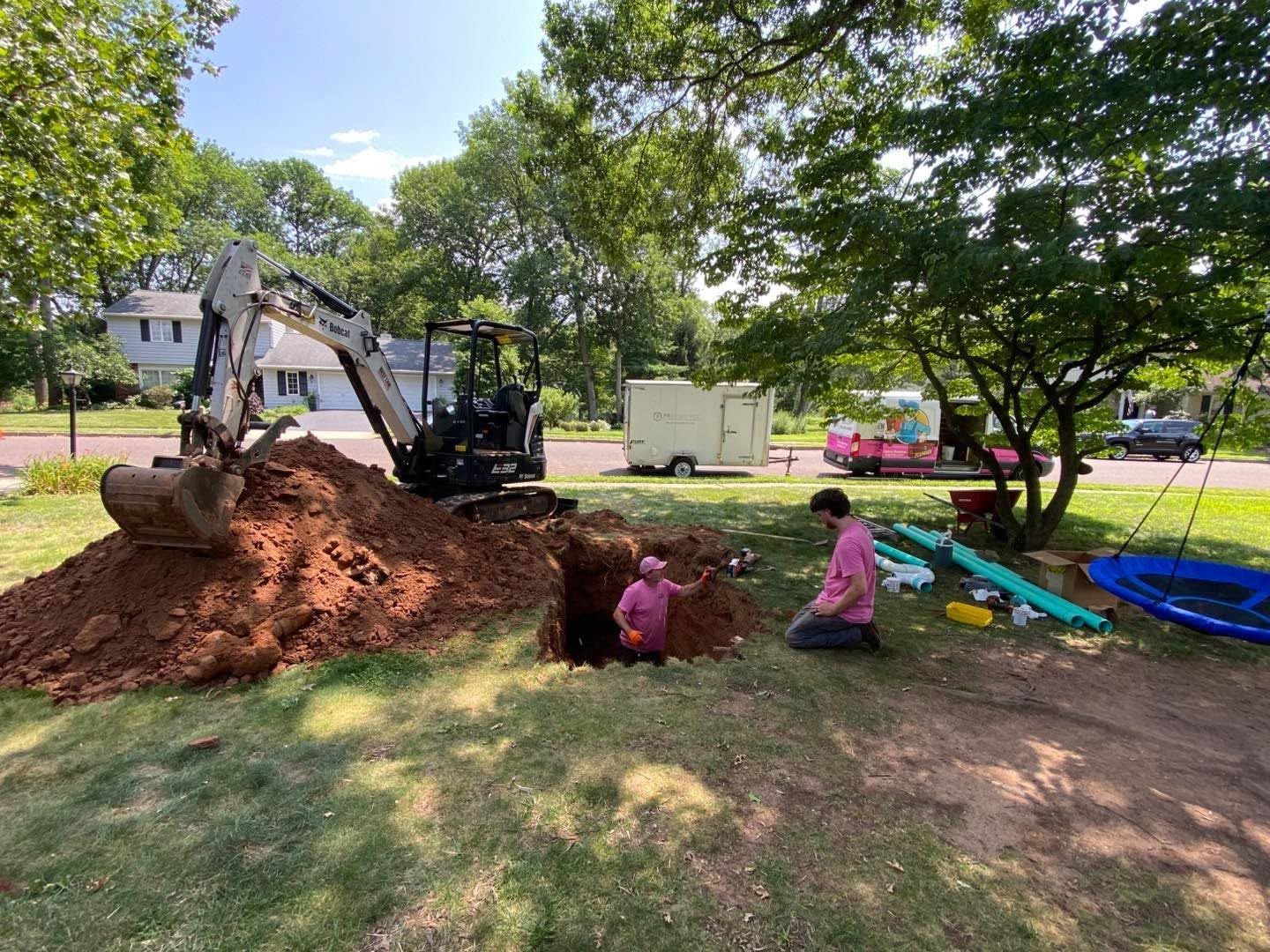 Excavator digging a large dirt pile in a grassy park while two people kneel nearby under trees