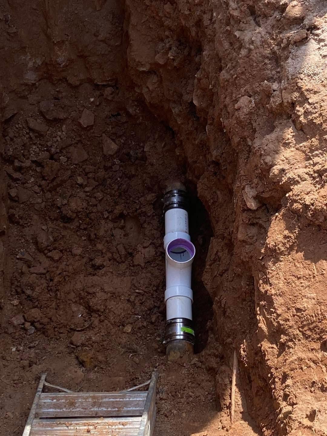 Exposed white PVC pipe fitting in a dirt trench beside a small wooden plank