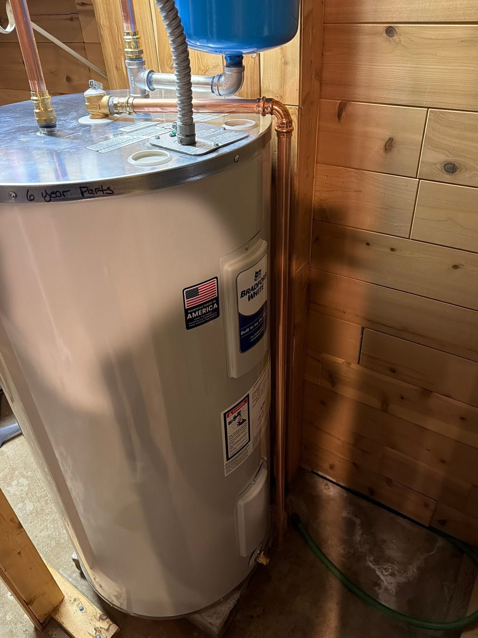 Large water heater in unfinished basement corner beside plywood wall and blue tank above it