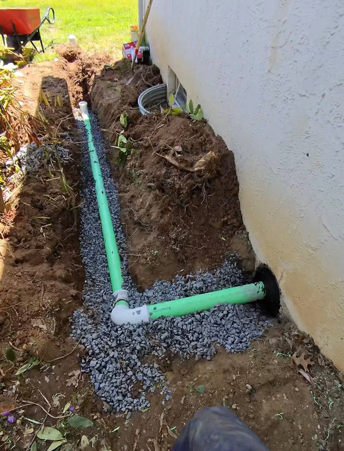 White PVC pipe trench beside house wall, with green elbow joints and gravel around an opening.