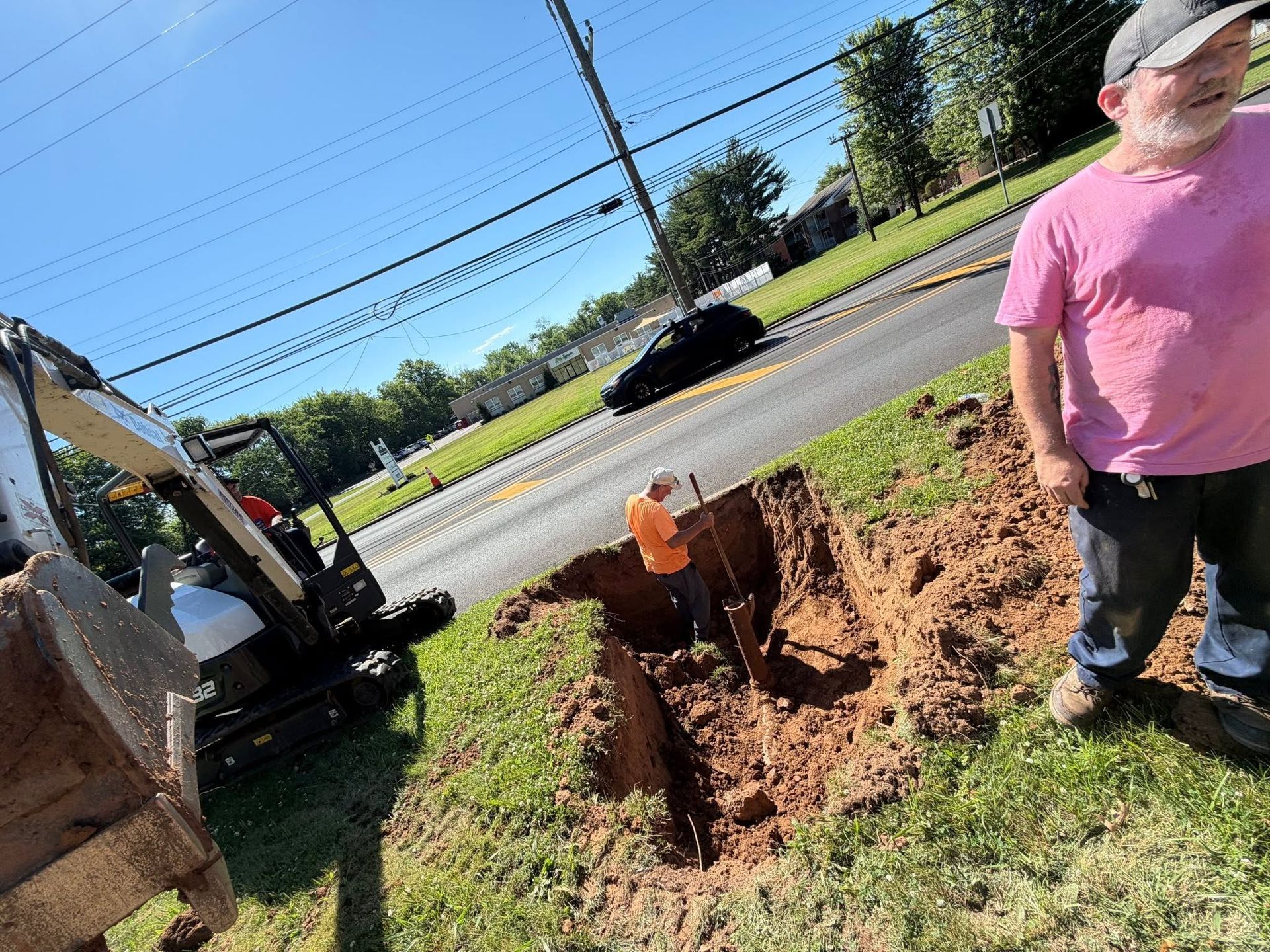 Workers dig a trench beside a road near a parked truck and a person in a pink shirt