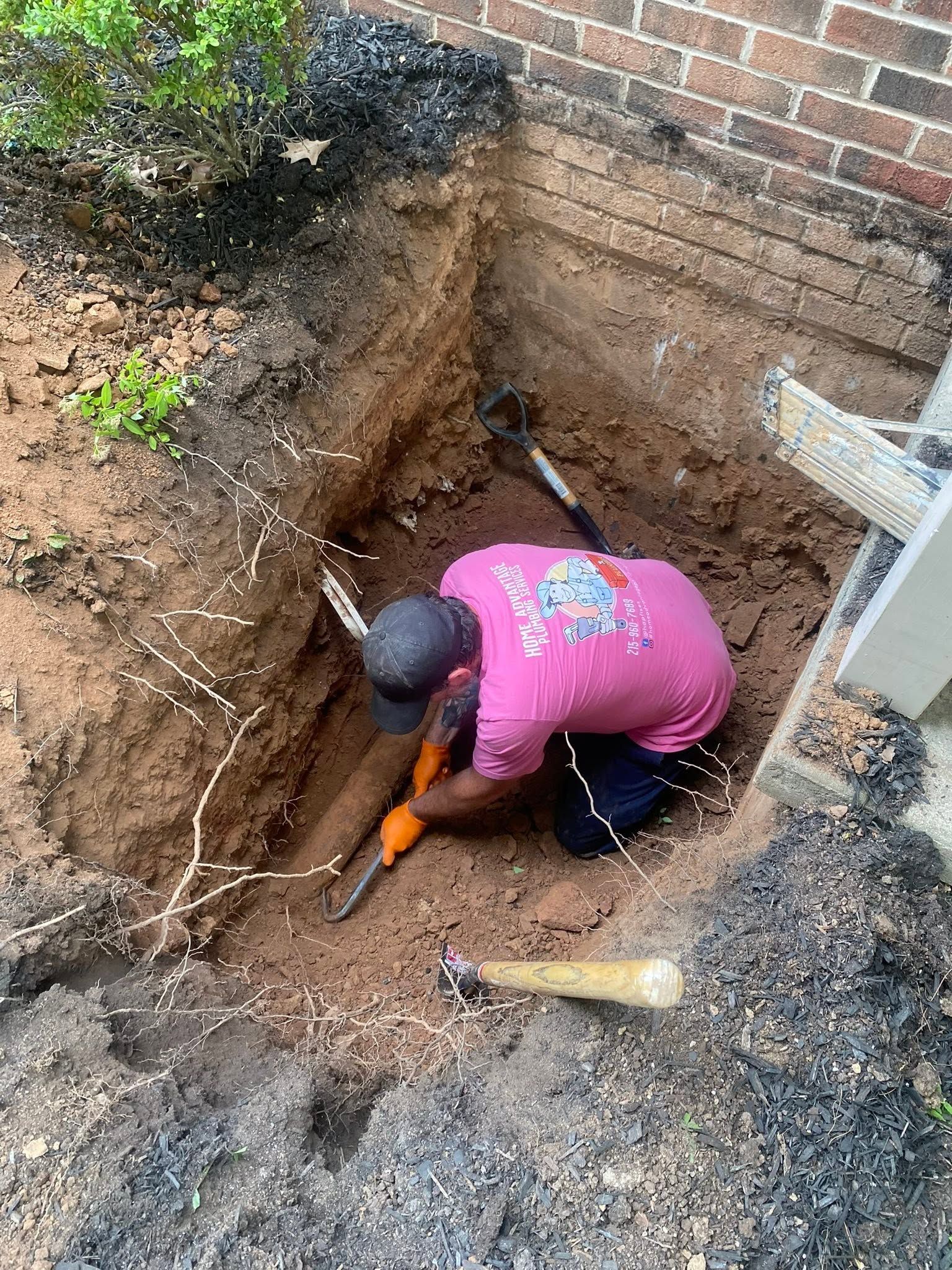 Person digging a trench beside a brick wall, using an orange shovel.