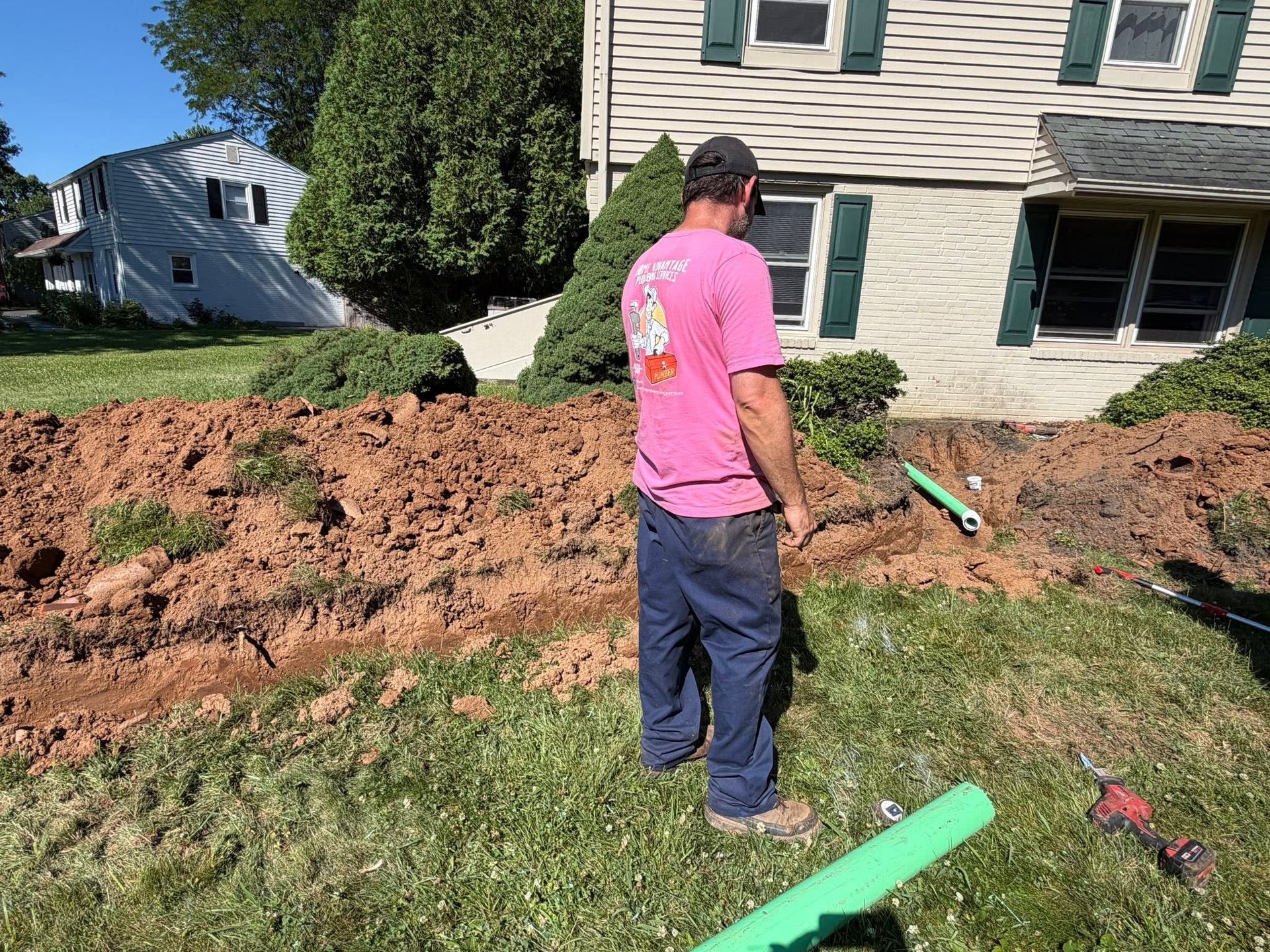 Person stands by a dug-up yard near a house, holding a shovel amid dirt and tools.