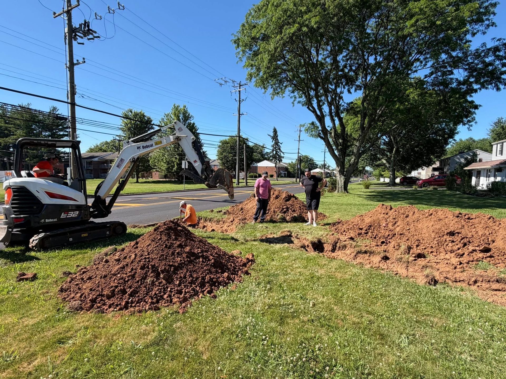 Roadwork crew with excavator and dirt piles beside a grassy roadside on a sunny day