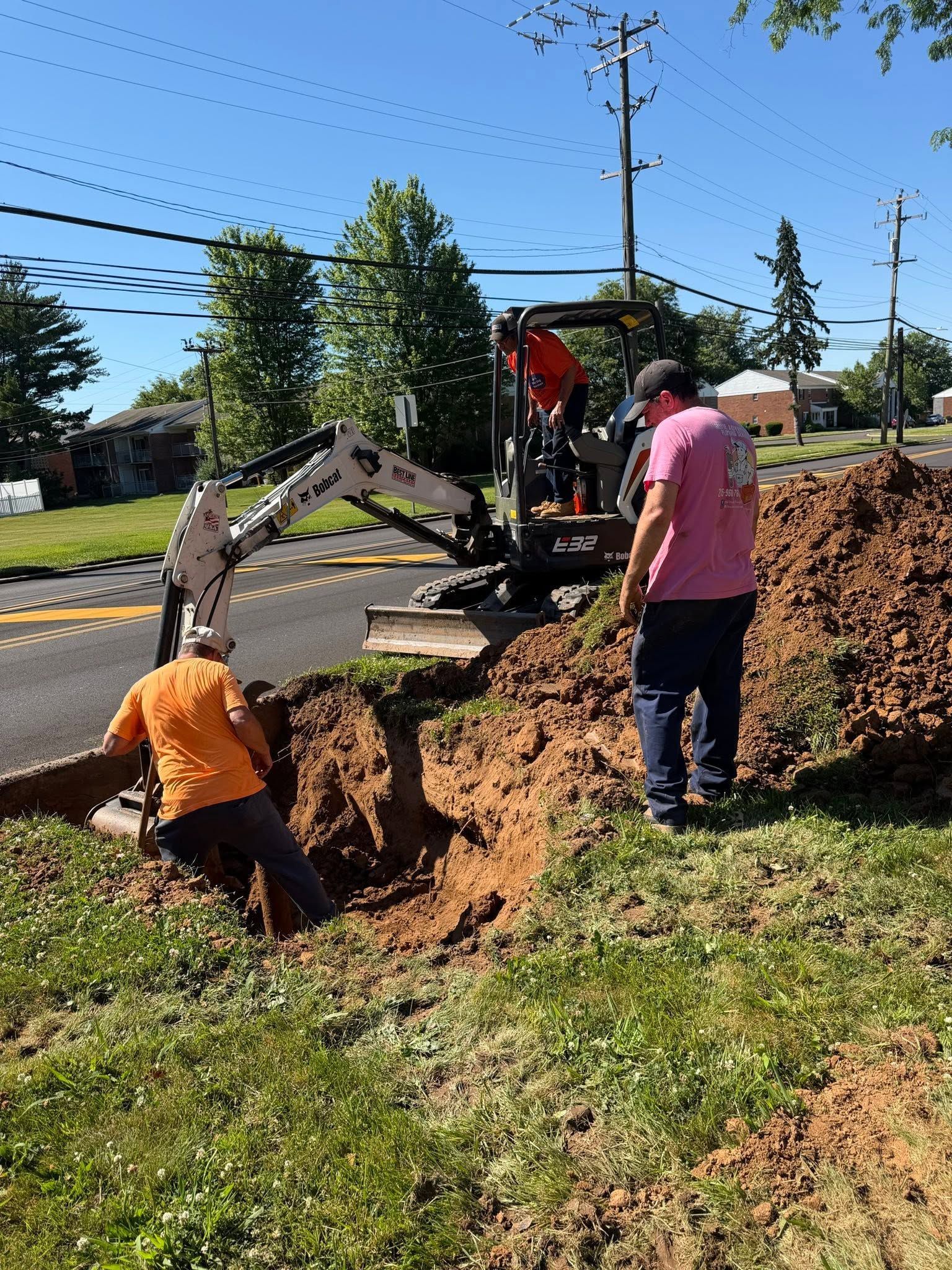 Workers dig a roadside trench beside an excavator on a sunny day.