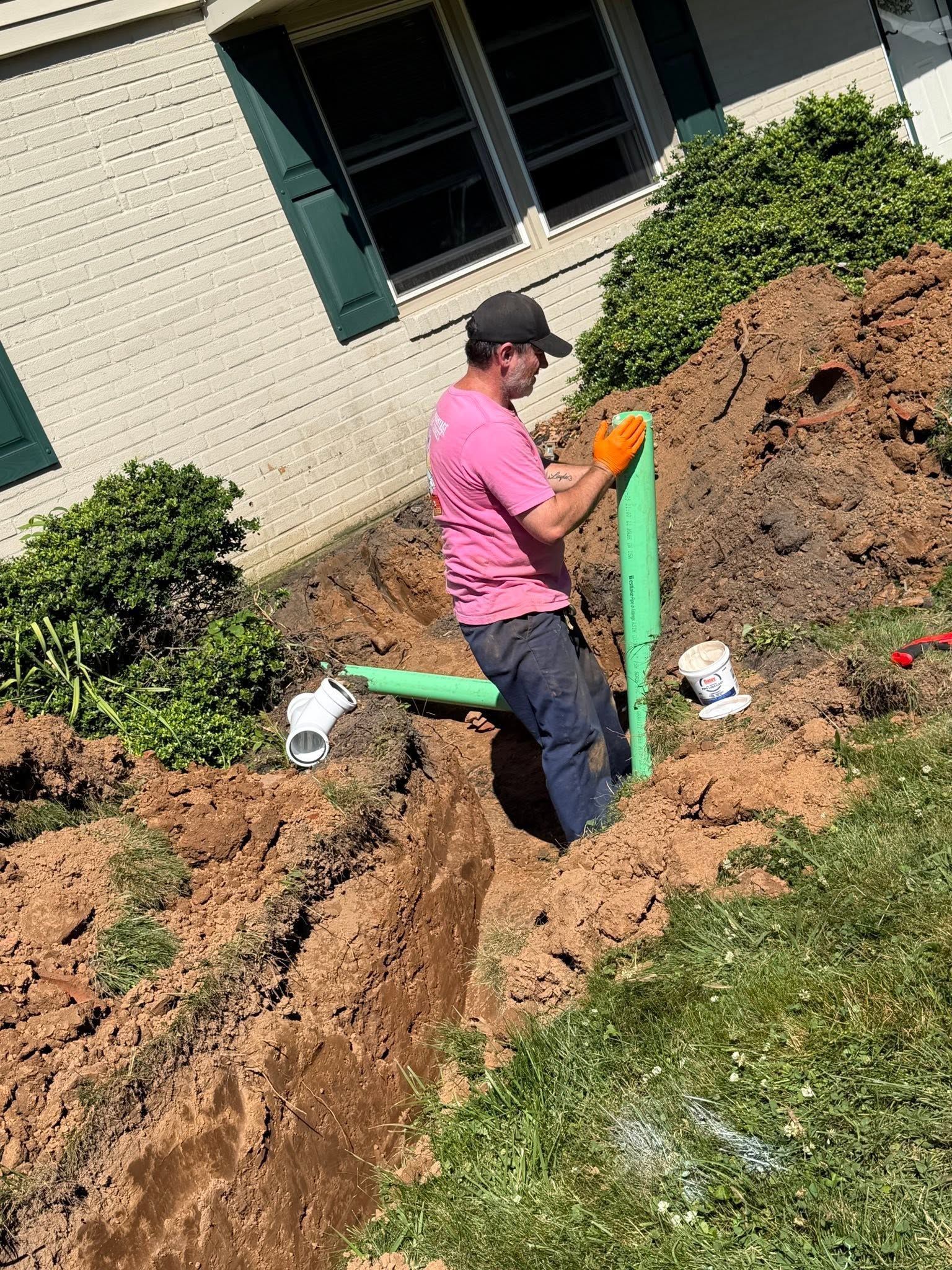 Person in a pink shirt working on a green pipe in a dirt trench beside a house.