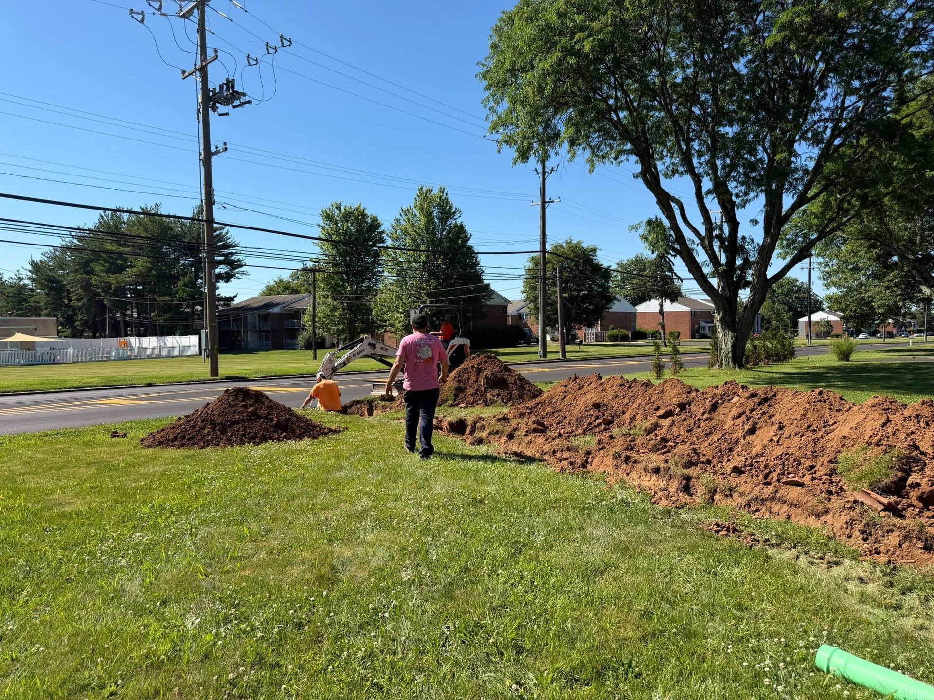 Workers in a grassy park dig a long trench beside a road on a sunny day