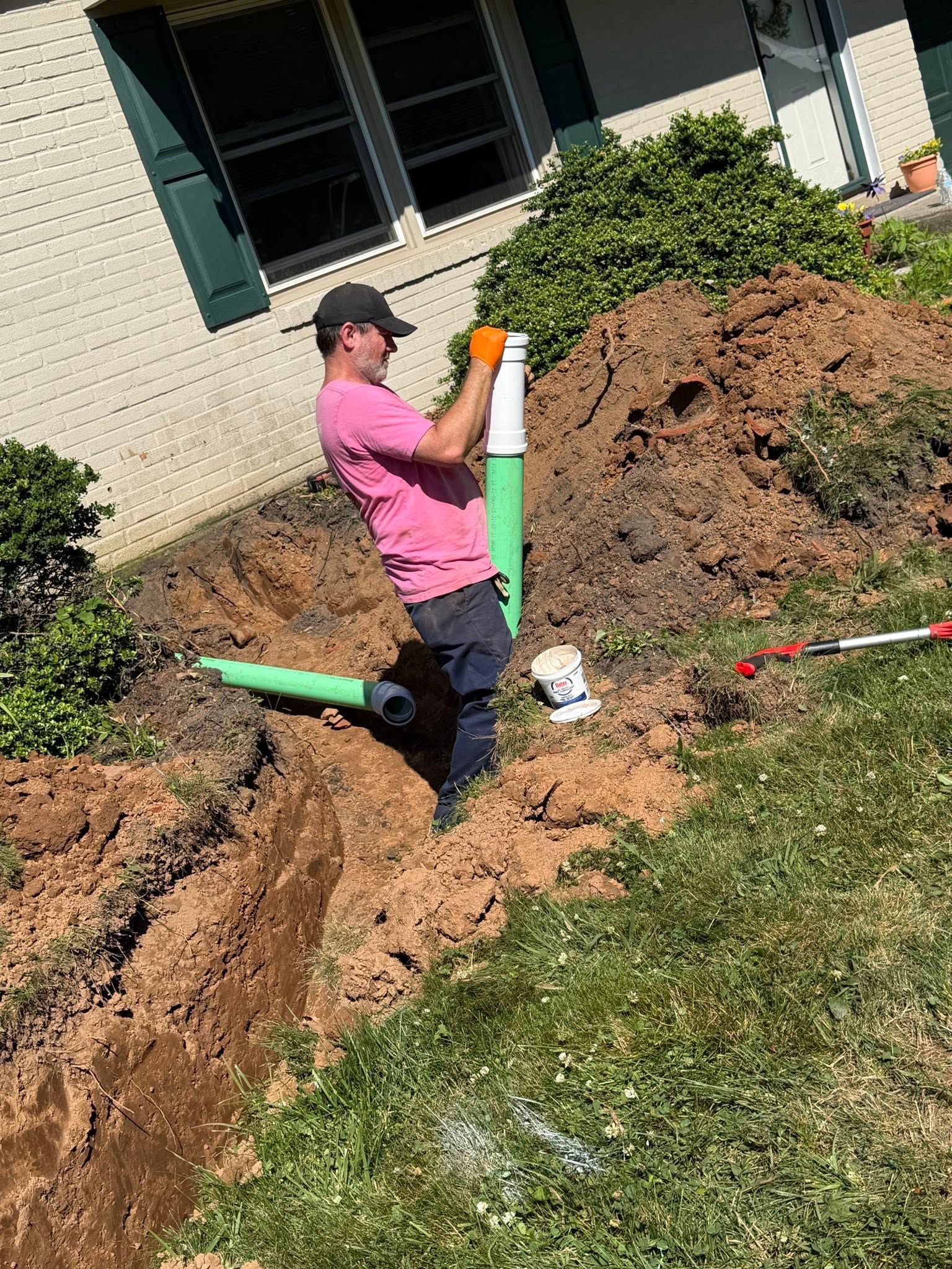 Person in a pink shirt standing in a trench beside green utility pipes during yard excavation work