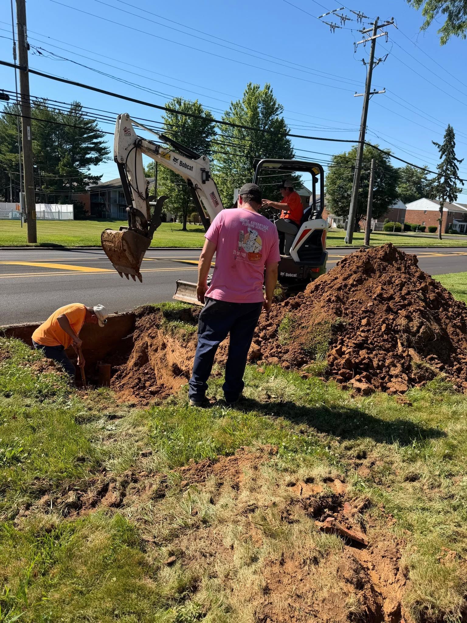 Workers digging a trench beside a road with an excavator and piles of dirt on a sunny day
