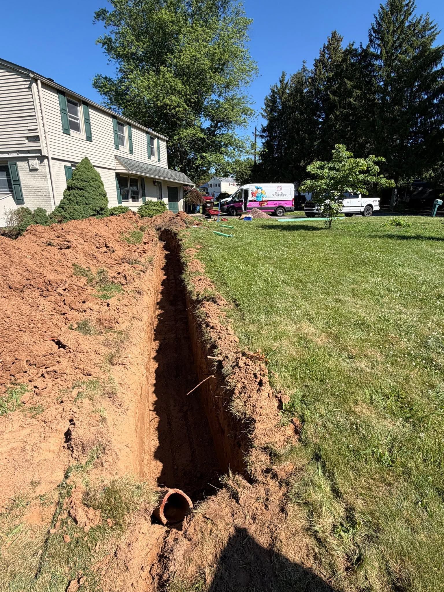 Deep trench dug beside a house in a grassy yard, with exposed pipe and utility vehicles in the background