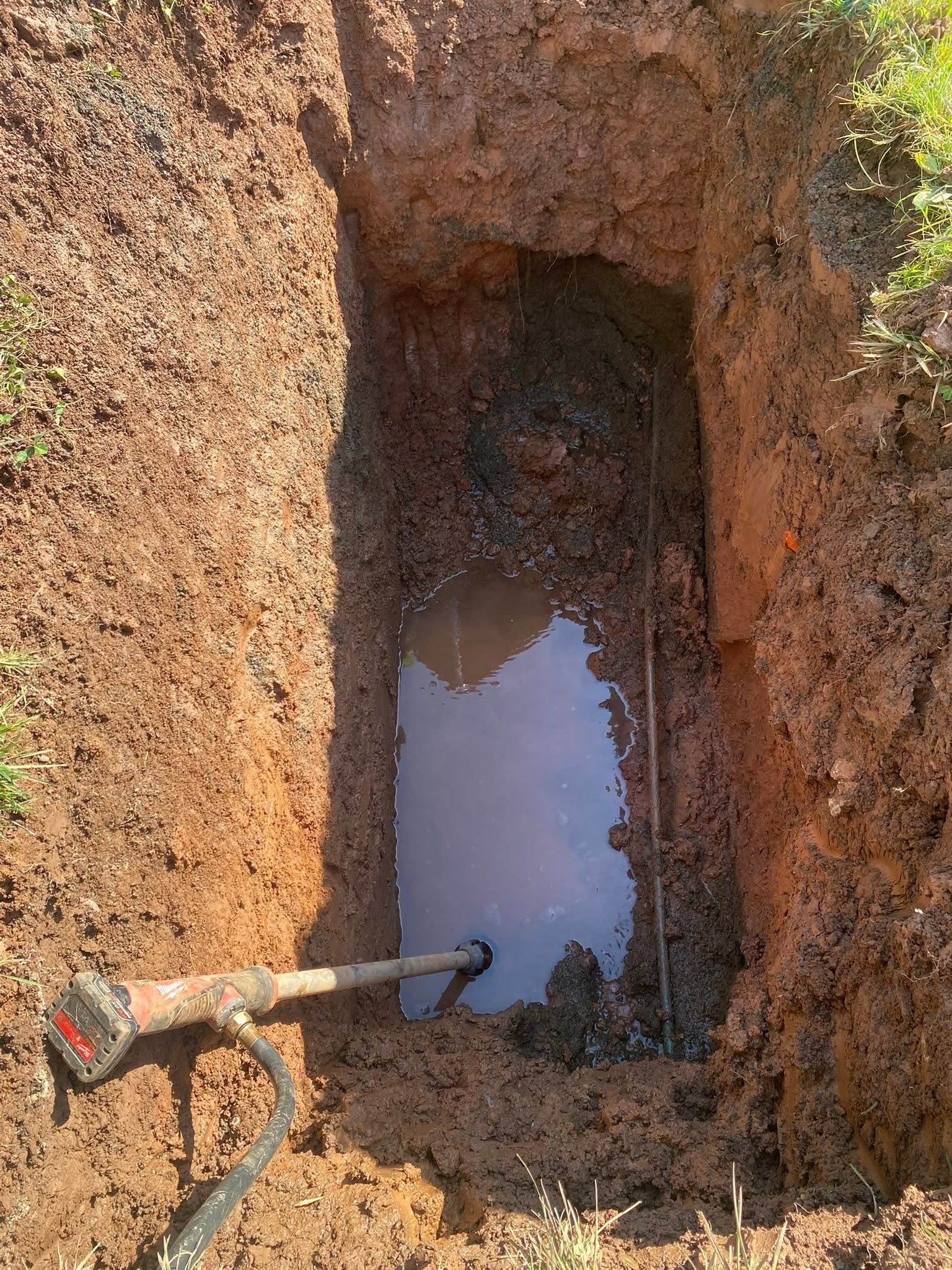 Narrow dirt trench with standing muddy water and a shovel handle at the edge