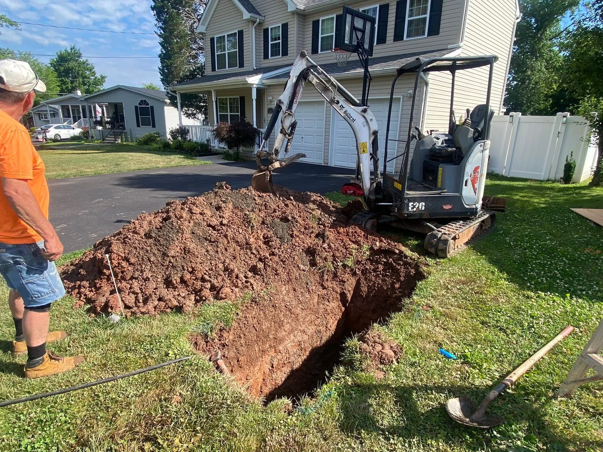 Excavator digging a trench beside a suburban house, with a large dirt mound in the driveway.