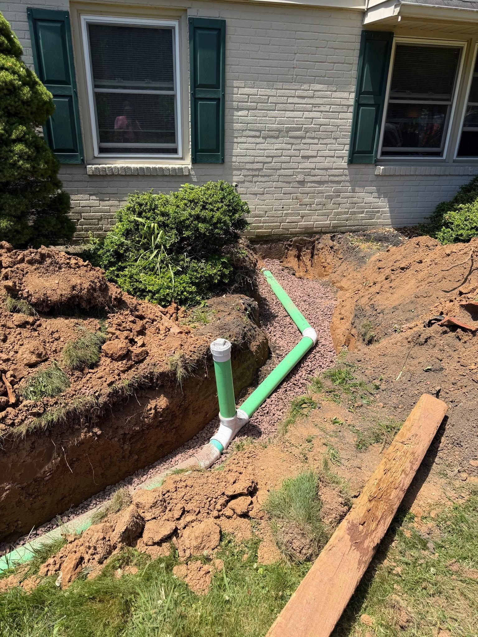 House exterior with a dug trench and exposed green PVC pipes beside the foundation.