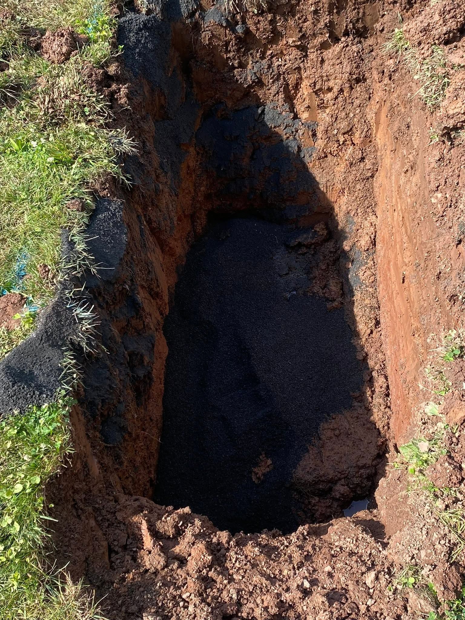 Deep rectangular hole in red soil with dark shadow and green grass along the edges