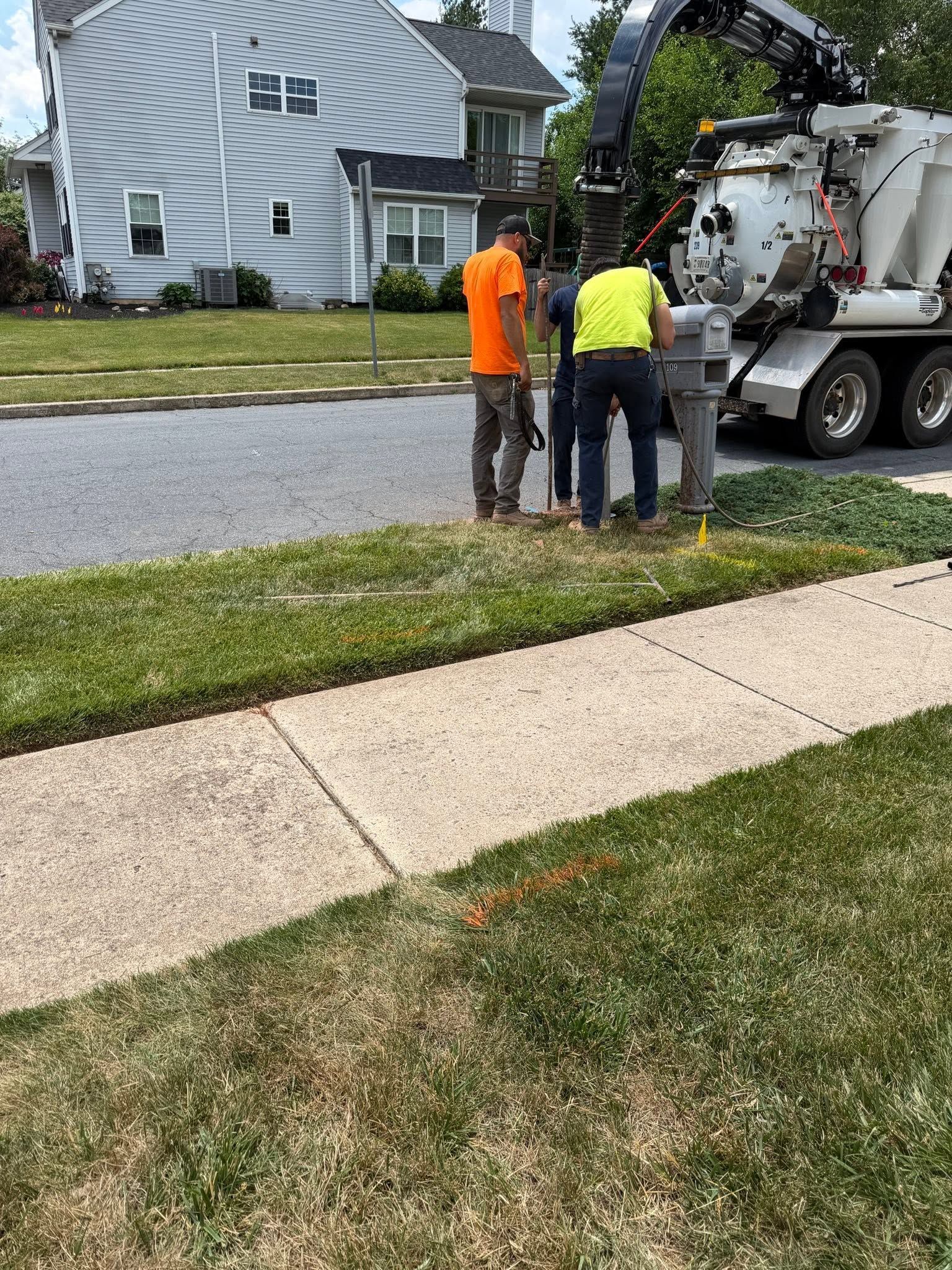 Two workers in orange and yellow vests beside a truck on a suburban street, near a house and sidewalk.