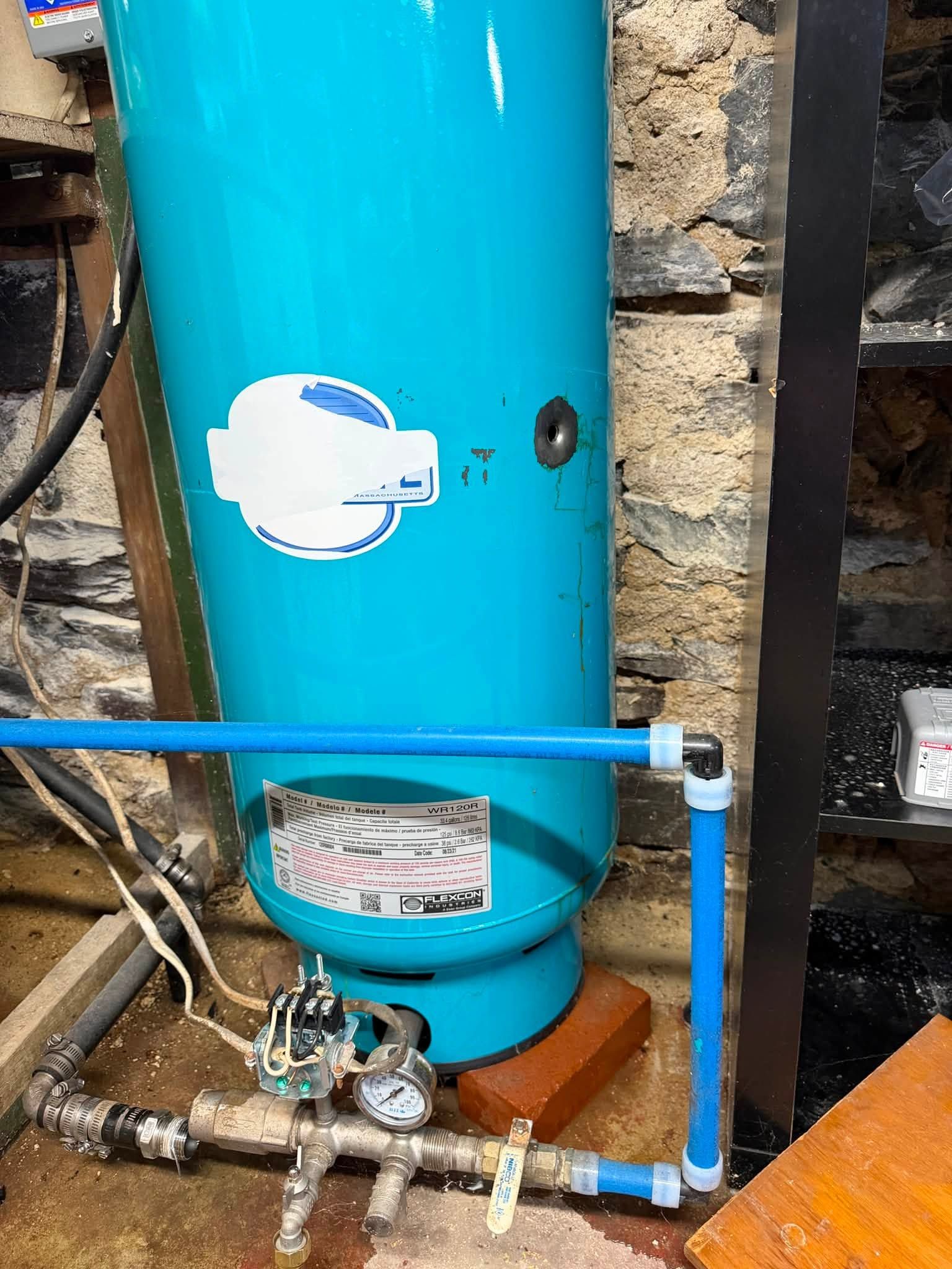 Blue water softener tank with pipes and valves in a utility area against a stone wall