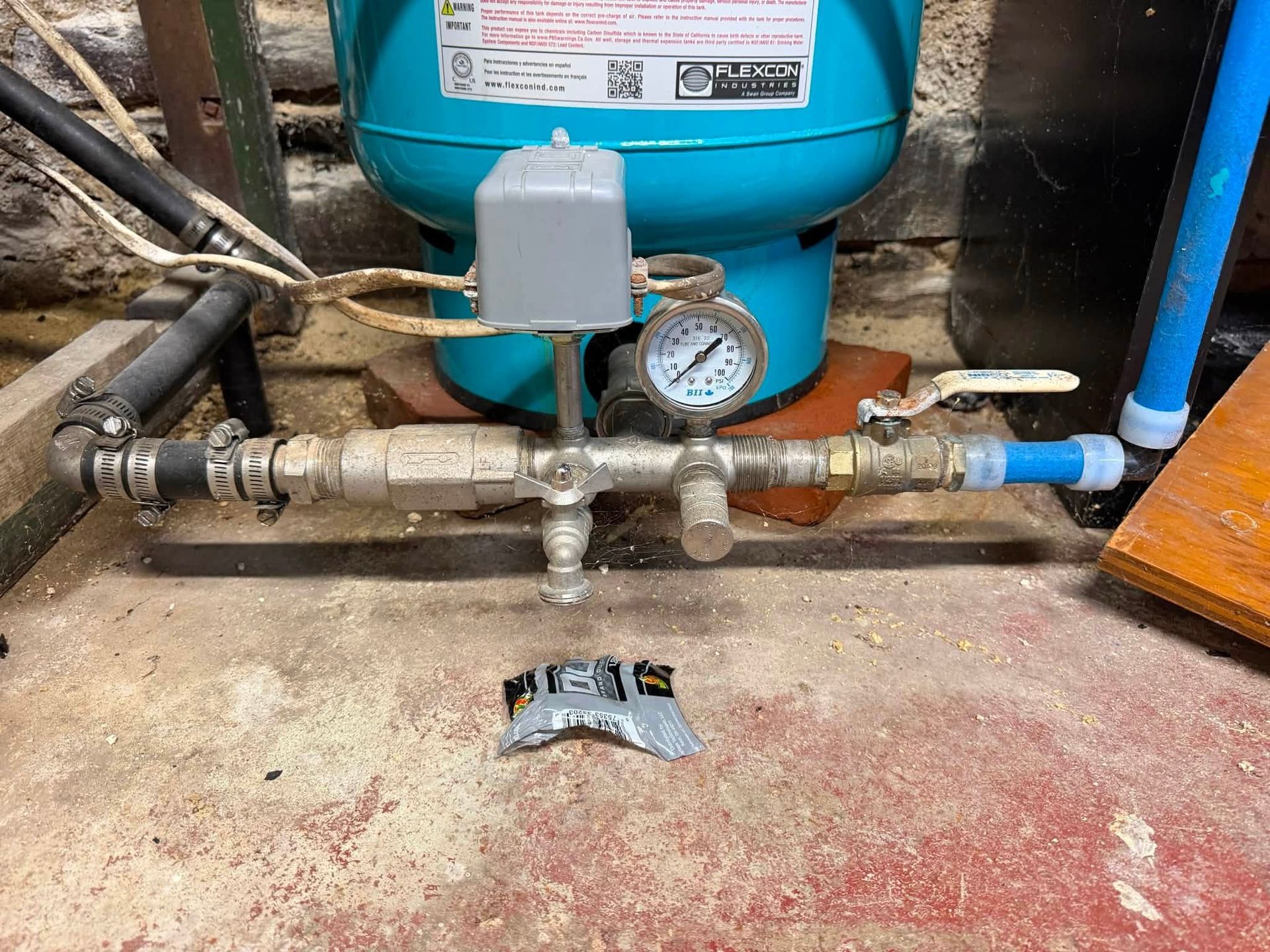 Water pressure tank and gauge with pipes and valves in a utility area, with a broken pipe fitting on the floor.
