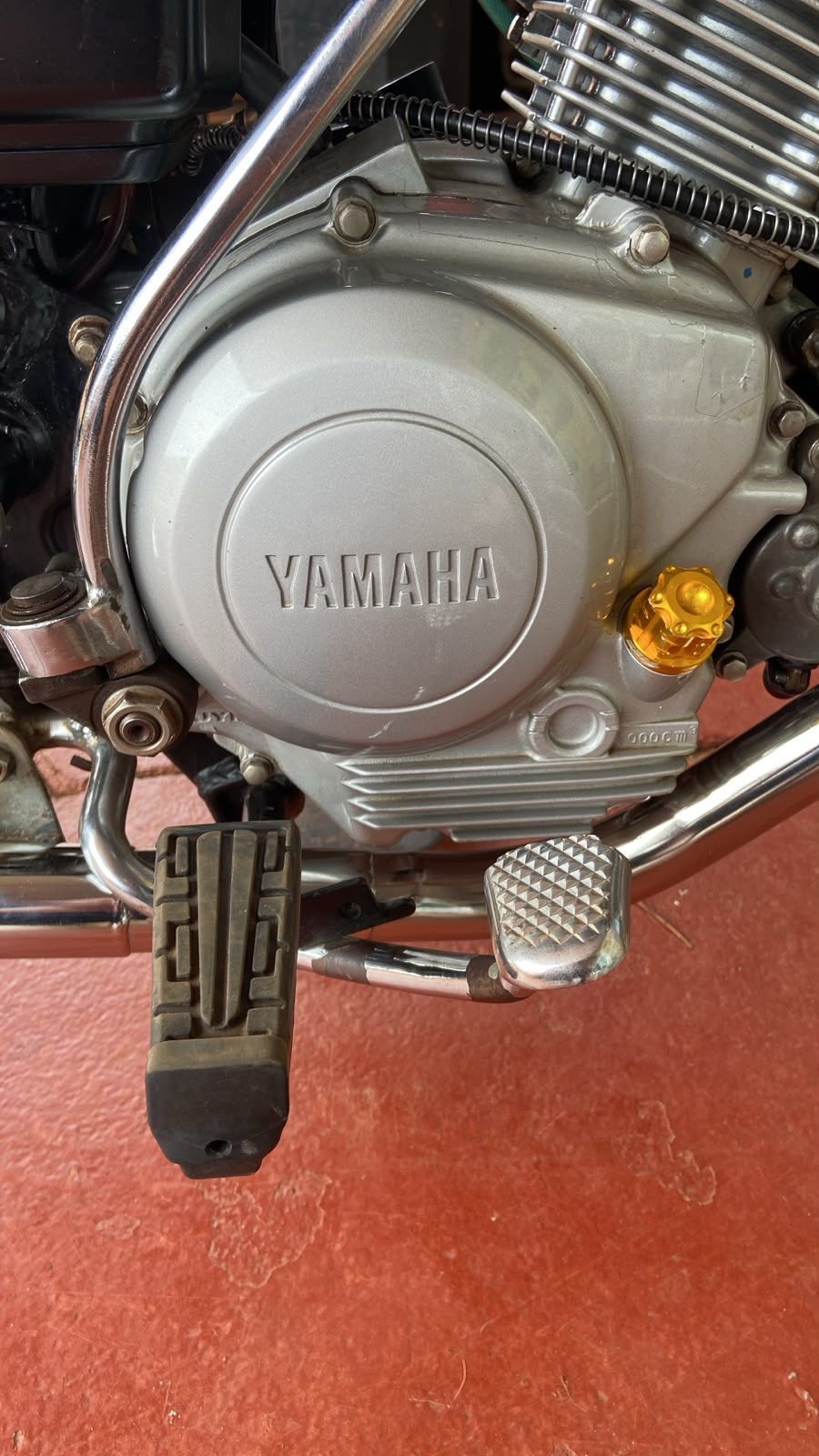 Primer plano del motor de una motocicleta Yamaha, con carcasa plateada, chasis negro y estribera sobre un suelo rojo