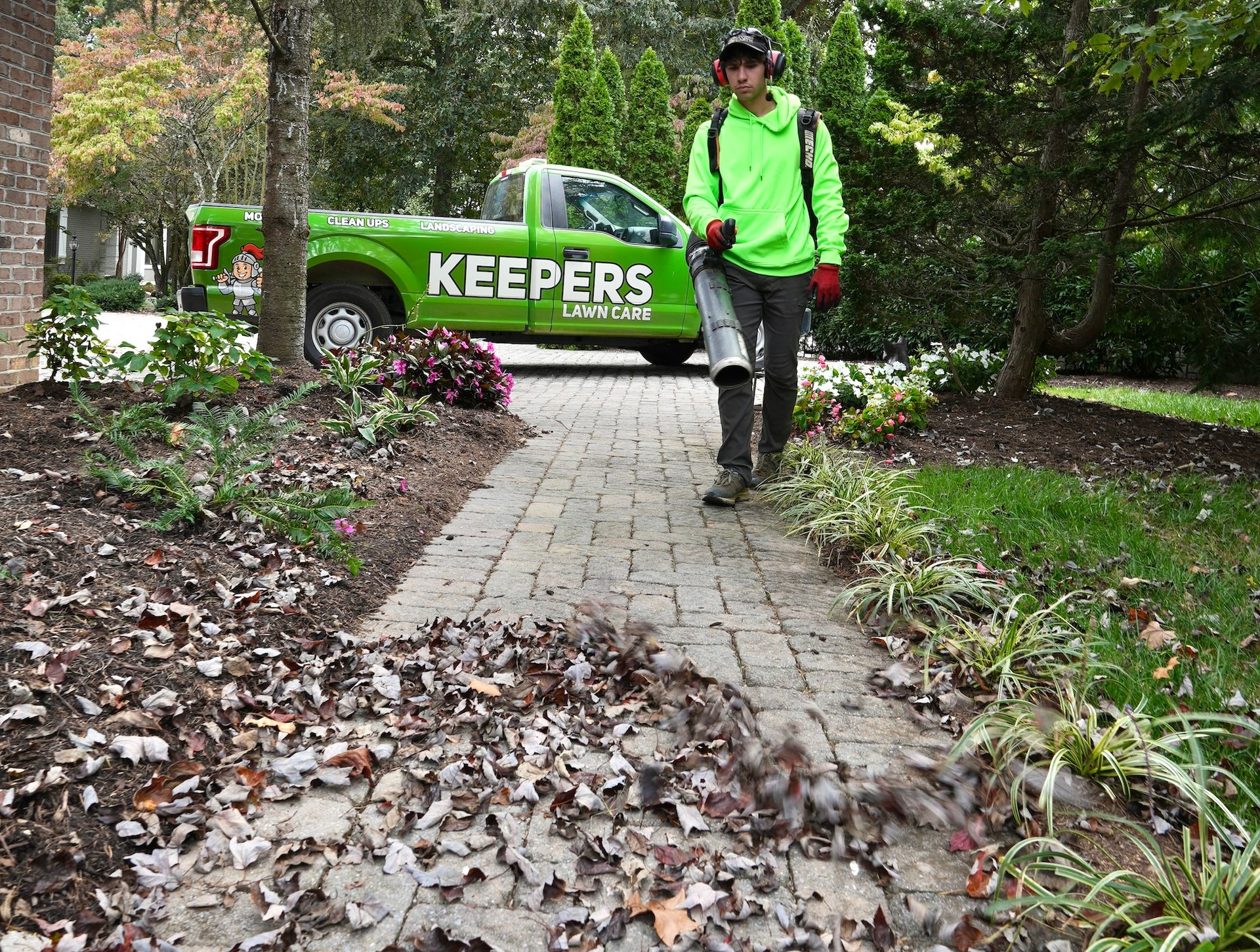 Person with backpack leaf blower, wearing ear protection, blowing leaves on a driveway in front of a house.