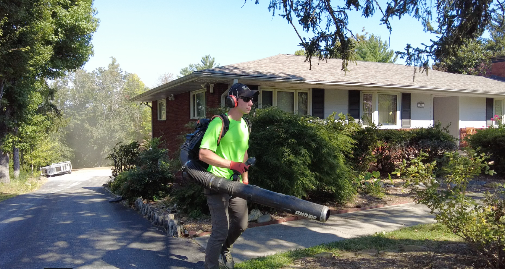 Person with backpack leaf blower, wearing ear protection, blowing leaves on a driveway in front of a house.