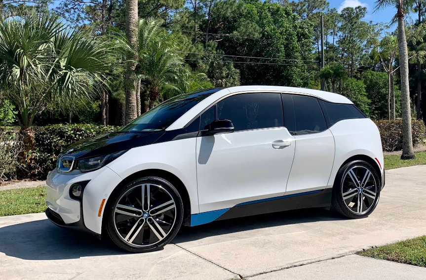 White and black BMW i3 electric car parked on a paved driveway with palm trees in the background.