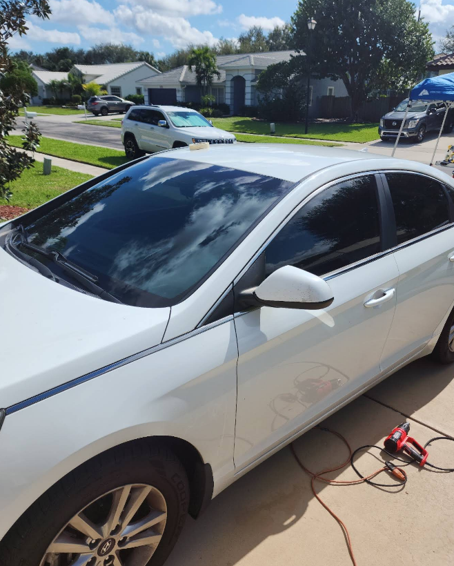 White car with tinted windows in a driveway, being worked on under a sunny sky in front of houses.
