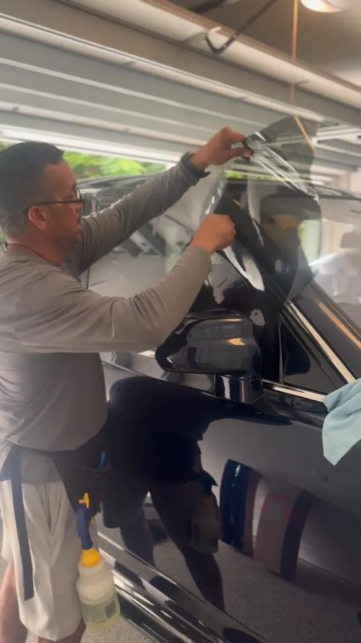 Man applying tint to a car window. He is in a garage, holding a film over the window.