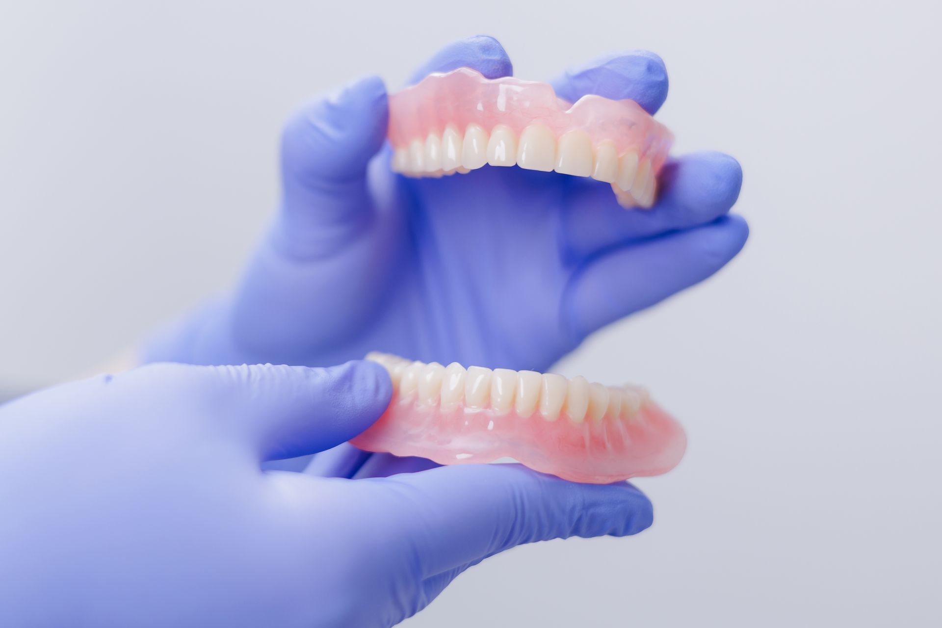 A close-up of a dental professional holding upper and lower dentures with purple gloves