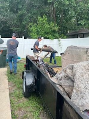 Three people loading junk into a trailer in a yard with a white fence.