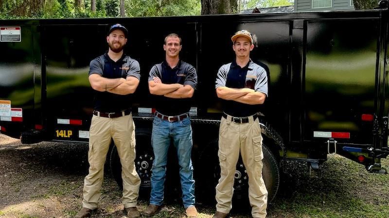 Three men in work attire stand in front of a black trailer with arms crossed.