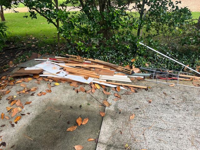 Pile of construction debris on concrete, next to bushes and grass. Fallen leaves scattered around.