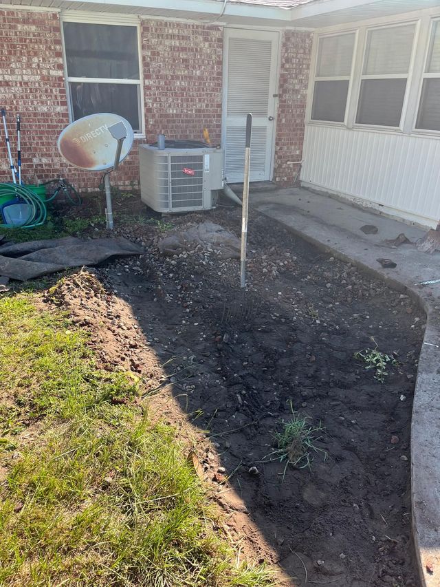 A newly dug garden bed with dark soil, next to a brick house and a concrete walkway.